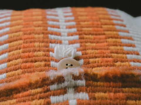 Close-up of a textured woven textile with a repeating orange, yellow, and white geometric pattern, featuring a small beige button on the front.