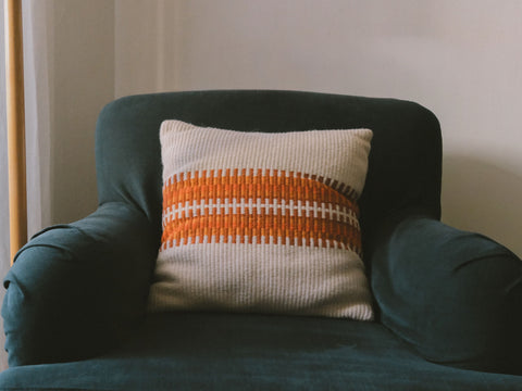 A square woven throw pillow with orange and white stripes, resting on a dark teal armchair against a neutral wall.
