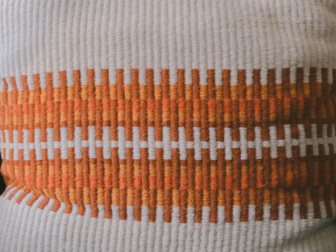 Close-up of a woven textile with a horizontal geometric stripe pattern in orange, brown, and white on a ribbed off-white background.